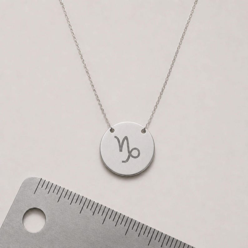 Pu&ograve; includere: Una collana color argento con un ciondolo circolare inciso con il simbolo zodiacale del Capricorno. Il ciondolo &egrave; posizionato accanto a un righello di metallo, che mostra che il disco circolare ha un diametro di circa 1,3 cm.