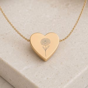 K&ouml;nnte beinhalten: Eine goldfarbene herzf&ouml;rmige Anh&auml;ngerkette mit einer zarten, d&uuml;nnen Kette und einer mittigen, lasergravierten Strichzeichnung einer einzelnen Chrysanthemenbl&uuml;te auf der glatten, polierten Oberfl&auml;che des Herzanh&auml;ngers.
