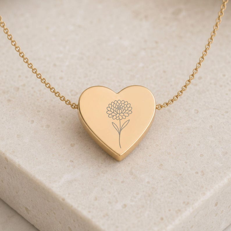 K&ouml;nnte beinhalten: Eine goldfarbene herzf&ouml;rmige Anh&auml;ngerkette mit einer zarten, d&uuml;nnen Kette und einer mittigen, lasergravierten Strichzeichnung einer einzelnen Chrysanthemenbl&uuml;te auf der glatten, polierten Oberfl&auml;che des Herzanh&auml;ngers.