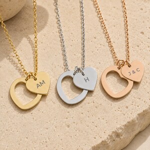 May include: Three necklaces featuring heart-shaped pendants on delicate chains. One gold necklace with AM, one silver necklace with H, and one rose gold necklace with J&C engraved on smaller heart charms attached to larger open heart frames.