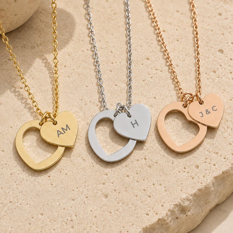 May include: Three necklaces featuring heart-shaped pendants on delicate chains. One gold necklace with AM, one silver necklace with H, and one rose gold necklace with J&C engraved on smaller heart charms attached to larger open heart frames.