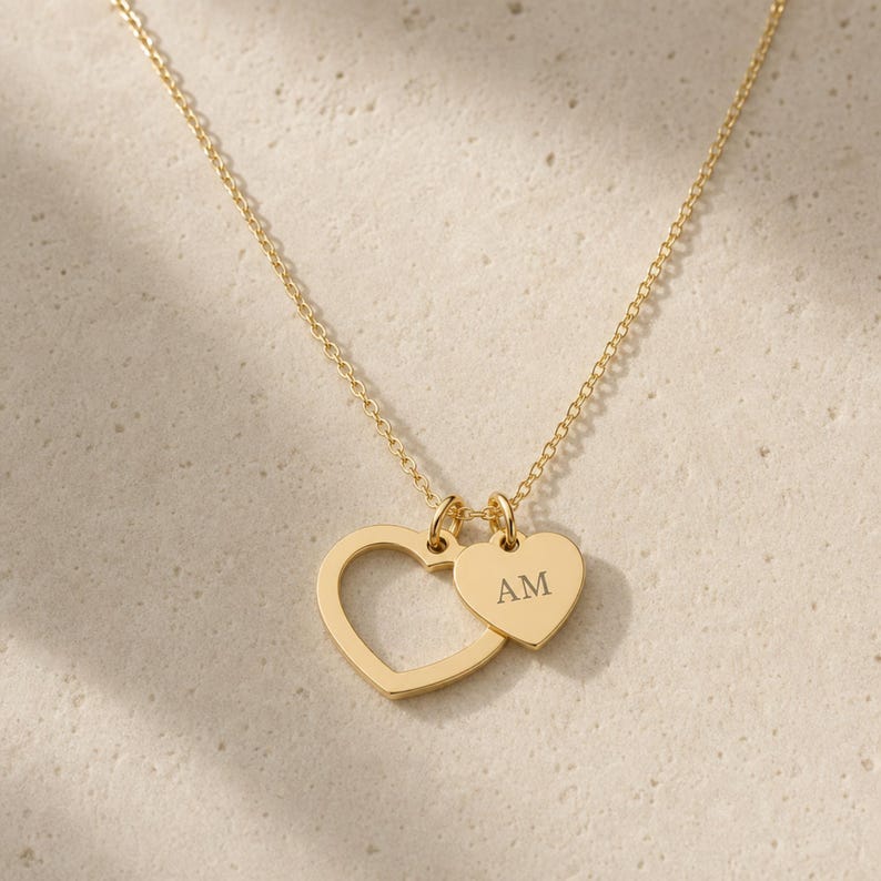 May include: A gold-toned necklace featuring a delicate chain and two heart-shaped pendants. One pendant is an open heart outline, while the smaller, solid heart pendant is engraved with the capital letters AM. The metal has a polished finish.