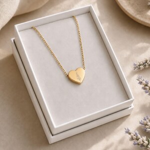 Puede incluir: Un collar con colgante en forma de coraz&oacute;n de tono dorado que presenta un delicado dise&ntilde;o floral grabado en la parte frontal. El collar se presenta dentro de una caja rectangular blanca, con la cadena extendi&eacute;ndose desde la parte superior.