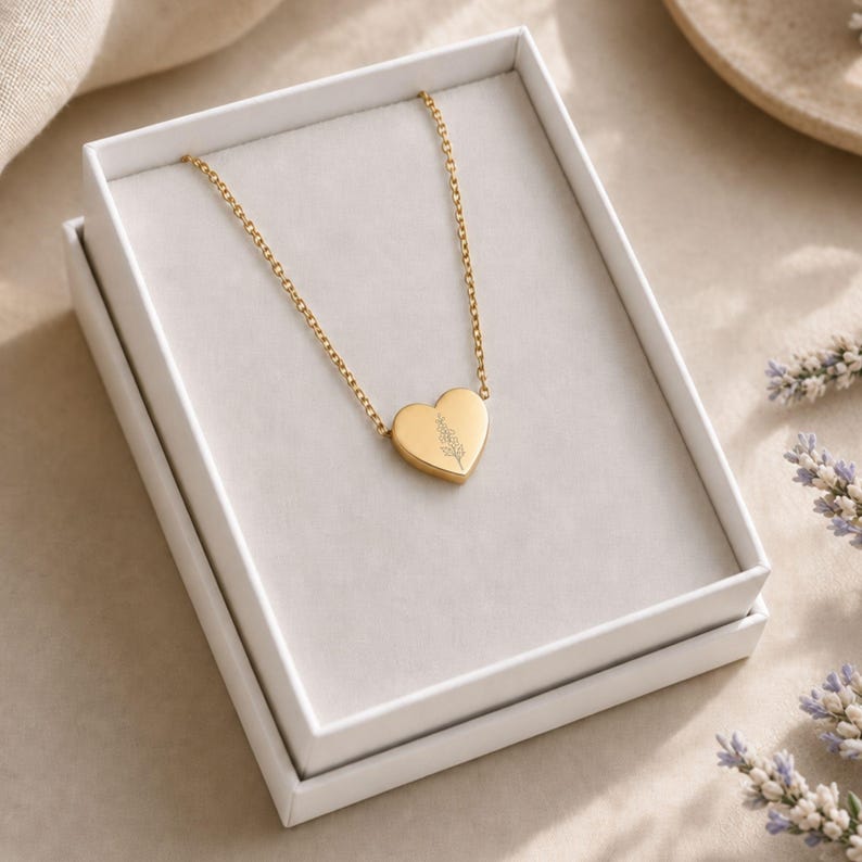 Puede incluir: Un collar con colgante en forma de coraz&oacute;n de tono dorado que presenta un delicado dise&ntilde;o floral grabado en la parte frontal. El collar se presenta dentro de una caja rectangular blanca, con la cadena extendi&eacute;ndose desde la parte superior.