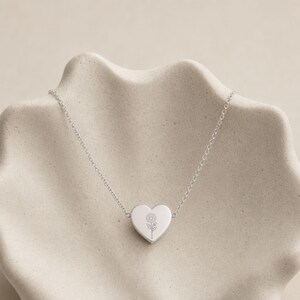 Pu&ograve; includere: Una collana color argento con una catenina sottile e un ciondolo centrale a forma di cuore. Il ciondolo &egrave; inciso con un'illustrazione a linee sottili di un singolo fiore di margherita centrato sulla superficie liscia e lucida.