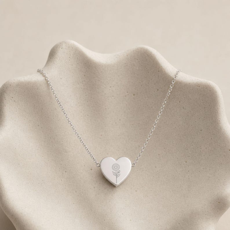 Pu&ograve; includere: Una collana color argento con una catenina sottile e un ciondolo centrale a forma di cuore. Il ciondolo &egrave; inciso con un'illustrazione a linee sottili di un singolo fiore di margherita centrato sulla superficie liscia e lucida.