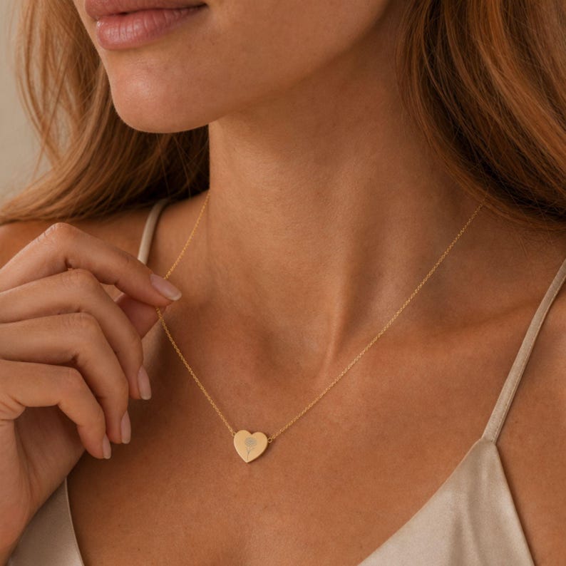 K&ouml;nnte beinhalten: Eine goldfarbene Halskette mit einer zarten Kette und einem kleinen, flachen herzf&ouml;rmigen Anh&auml;nger. In der Mitte des Herzanh&auml;ngers befindet sich ein dezentes, eingraviertes Blumenmuster. Die Kette liegt am Hals an und hat ein schlichtes Finish.