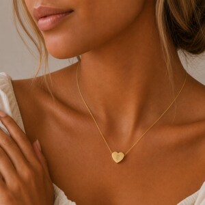 May include: A gold-toned necklace featuring a delicate heart-shaped pendant. The pendant is centered on a thin chain and displays a small, engraved floral design on its surface. The metal has a smooth, polished finish that reflects light.