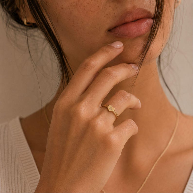 Puede incluir: Un anillo de oro con un peque&ntilde;o dije plano en forma de coraz&oacute;n centrado en una banda fina y pulida. El dije de coraz&oacute;n tiene una superficie lisa y reflectante, unido a un delicado anillo de metal circular que se ajusta al dedo.