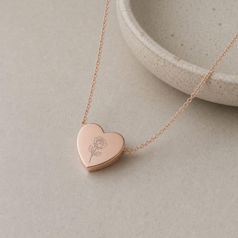 May include: A rose gold heart-shaped pendant necklace featuring a delicate, fine-line engraved rose flower on the front surface. The pendant is suspended from a matching rose gold chain with small, uniform circular links, creating a simple design.