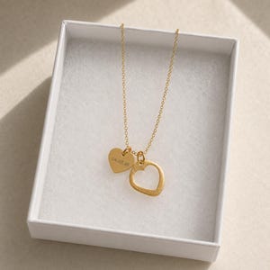 May include: A gold-colored necklace featuring a delicate chain with two heart-shaped pendants. One pendant is a solid heart engraved with the date 04.03.26, while the other is an open heart outline. The necklace is presented inside a white box.