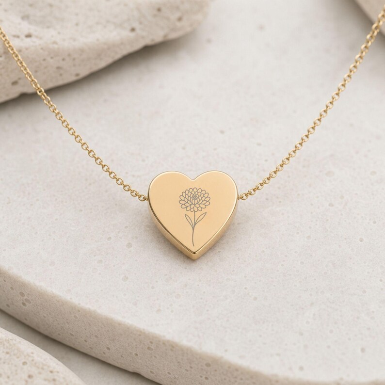 K&ouml;nnte beinhalten: Eine goldfarbene herzf&ouml;rmige Anh&auml;ngerkette mit einer feinen Kette und einer mittigen Gravur einer einzelnen Blume mit einer runden, texturierten Bl&uuml;te und einem d&uuml;nnen Stiel. Die Metalloberfl&auml;che ist glatt und poliert mit einem warmen Glanz.