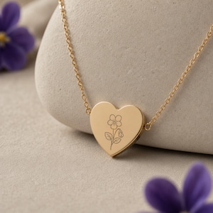 Puede incluir: Un collar con colgante en forma de coraz&oacute;n de tono dorado que presenta un delicado grabado de l&iacute;neas de una flor solitaria con hojas. El colgante est&aacute; unido a una fina cadena dorada, creando una pieza de joyer&iacute;a sencilla para el uso diario.