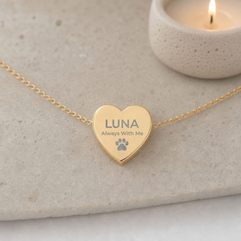May include: A gold-toned heart-shaped pendant necklace featuring the engraved text LUNA, the phrase Always With Me, and a small paw print icon. The pendant is attached to a delicate gold-toned chain, creating a simple and elegant piece of jewelry.
