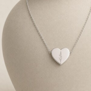 May include: A silver-toned heart-shaped pendant necklace featuring a delicate, fine-line engraved floral design on the front surface. The pendant is suspended from a thin, matching silver-toned metal chain with small, uniform circular links.