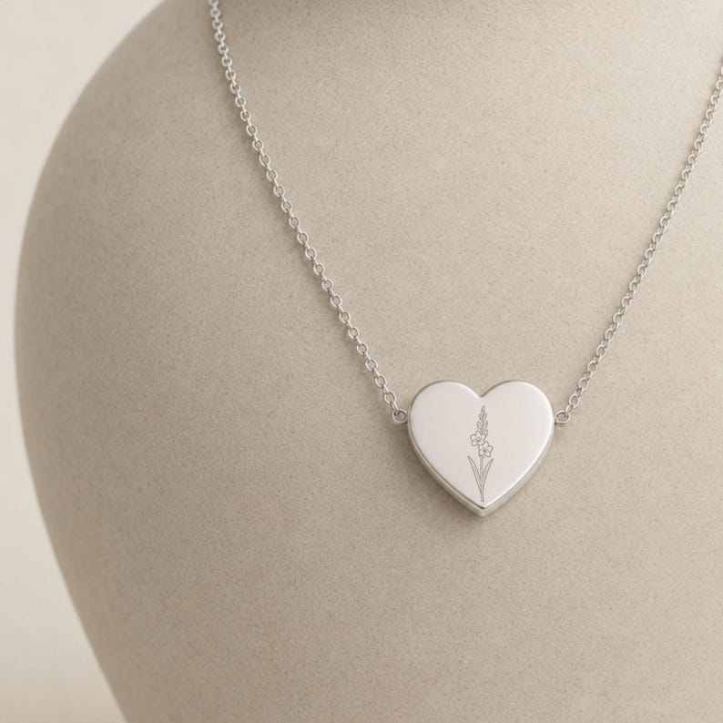 May include: A silver-toned heart-shaped pendant necklace featuring a delicate, fine-line engraved floral design on the front surface. The pendant is suspended from a thin, matching silver-toned metal chain with small, uniform circular links.