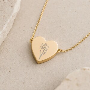 May include: A gold-toned heart-shaped pendant necklace featuring a delicate, dark-lined engraving of a single daffodil flower centered on the front surface. The pendant is attached to a thin, matching gold-toned metal chain link necklace.