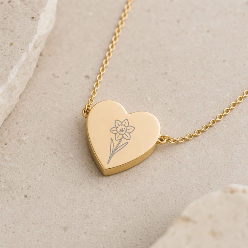 May include: A gold-toned heart-shaped pendant necklace featuring a delicate, dark-lined engraving of a single daffodil flower centered on the front surface. The pendant is attached to a thin, matching gold-toned metal chain link necklace.