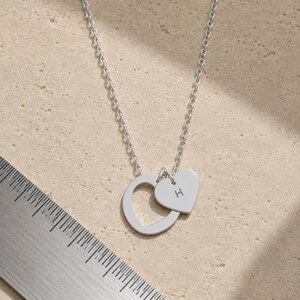 May include: A silver-toned necklace featuring a circular pendant and a smaller heart-shaped charm engraved with the letter H. A metal ruler is visible in the corner, showing the pendant assembly measures approximately 0.75 inches in total width.