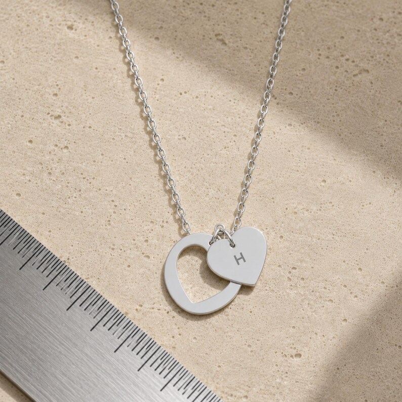 May include: A silver-toned necklace featuring a circular pendant and a smaller heart-shaped charm engraved with the letter H. A metal ruler is visible in the corner, showing the pendant assembly measures approximately 0.75 inches in total width.