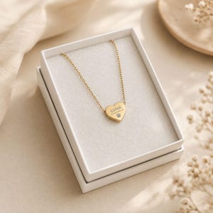 May include: A gold-toned metal necklace featuring a heart-shaped pendant. The pendant is engraved with the name LUNA, the phrase Always With Me, and a small paw print icon. The necklace is presented inside a white rectangular gift box.