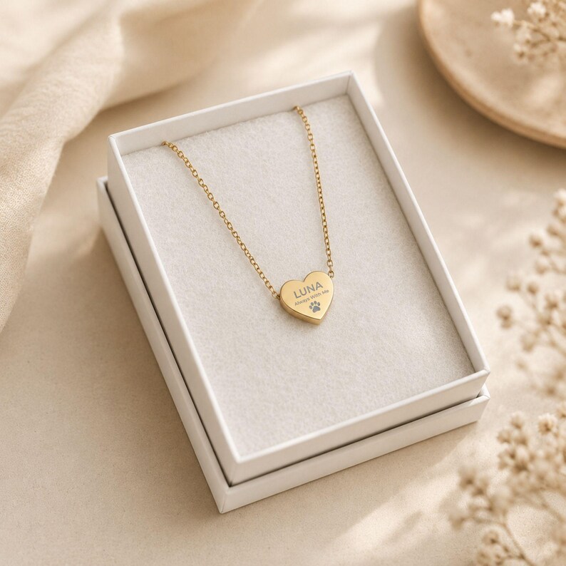 May include: A gold-toned metal necklace featuring a heart-shaped pendant. The pendant is engraved with the name LUNA, the phrase Always With Me, and a small paw print icon. The necklace is presented inside a white rectangular gift box.