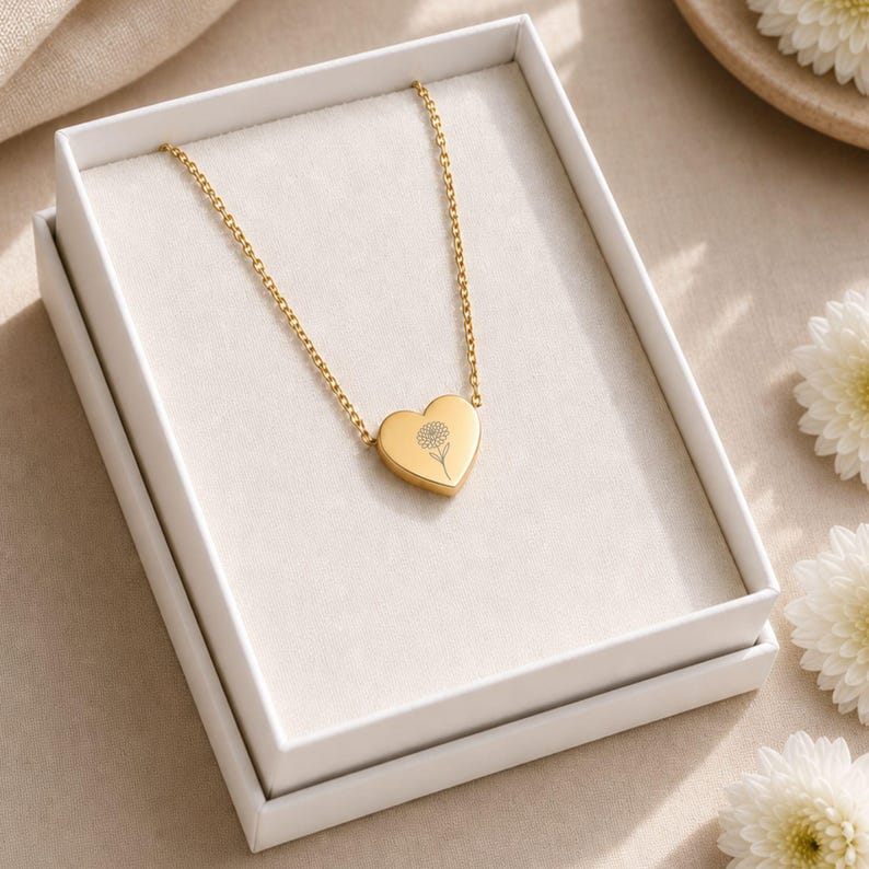 K&ouml;nnte beinhalten: Eine goldfarbene Halskette mit herzf&ouml;rmigem Anh&auml;nger, der auf der Vorderseite eine feine Strichzeichnung einer Blume aufweist. Die Kette liegt in einer wei&szlig;en, rechteckigen Geschenkbox mit einem hellen Stofffutter.