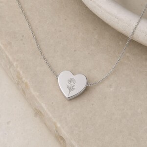 May include: A silver-toned heart-shaped pendant necklace featuring a delicate, fine-line engraving of a single flower on the front surface. The pendant is attached to a thin, matching silver-toned chain that extends from both sides of the heart.