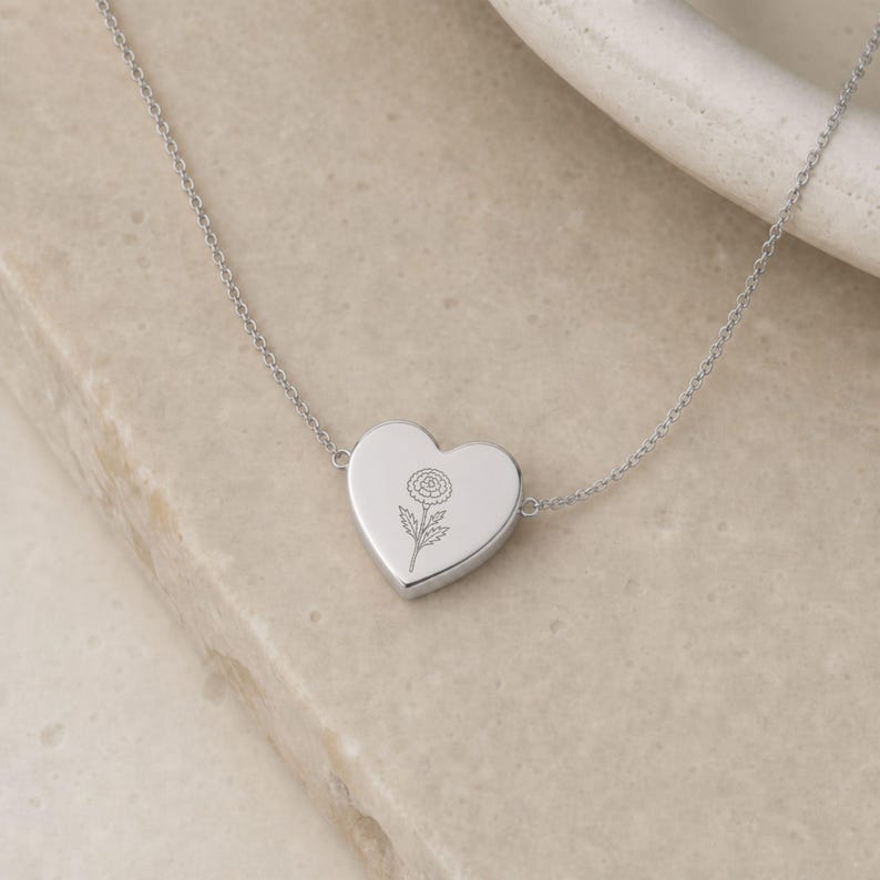 May include: A silver-toned heart-shaped pendant necklace featuring a delicate, fine-line engraving of a single flower on the front surface. The pendant is attached to a thin, matching silver-toned chain that extends from both sides of the heart.