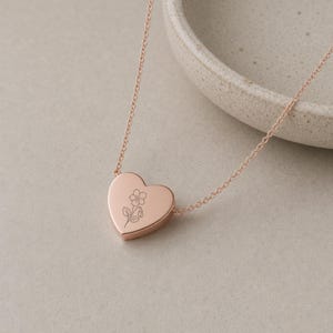Puede incluir: Un collar con colgante en forma de coraz&oacute;n de oro rosa que presenta una cadena fina y delicada con una superficie lisa y pulida. El centro del coraz&oacute;n est&aacute; grabado con una ilustraci&oacute;n sencilla de l&iacute;neas finas de una flor con tallo.