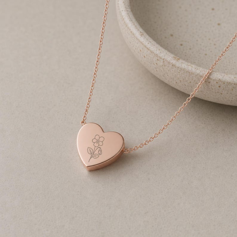 Puede incluir: Un collar con colgante en forma de coraz&oacute;n de oro rosa que presenta una cadena fina y delicada con una superficie lisa y pulida. El centro del coraz&oacute;n est&aacute; grabado con una ilustraci&oacute;n sencilla de l&iacute;neas finas de una flor con tallo.