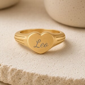 Puede incluir: Un anillo de tono dorado con una cara en forma de coraz&oacute;n que tiene el nombre Leo grabado en letra cursiva. La banda del anillo muestra tres l&iacute;neas horizontales grabadas a cada lado del coraz&oacute;n, aportando un detalle texturizado al metal.