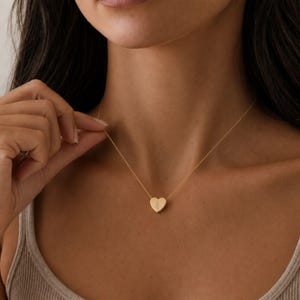 Puede incluir: Un collar de tono dorado que presenta un peque&ntilde;o colgante s&oacute;lido en forma de coraz&oacute;n suspendido de una cadena fina y delicada. El colgante de coraz&oacute;n tiene una superficie lisa y pulida con un sutil detalle de l&iacute;nea grabada vertical.