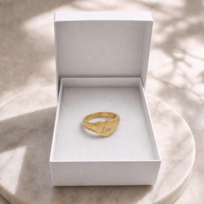 Puede incluir: Un anillo de sello dorado en forma de coraz&oacute;n con el nombre Leo grabado en letra cursiva, presentado dentro de un joyero rectangular blanco abierto con un forro interior suave, que muestra un acabado de metal pulido y liso en la banda.