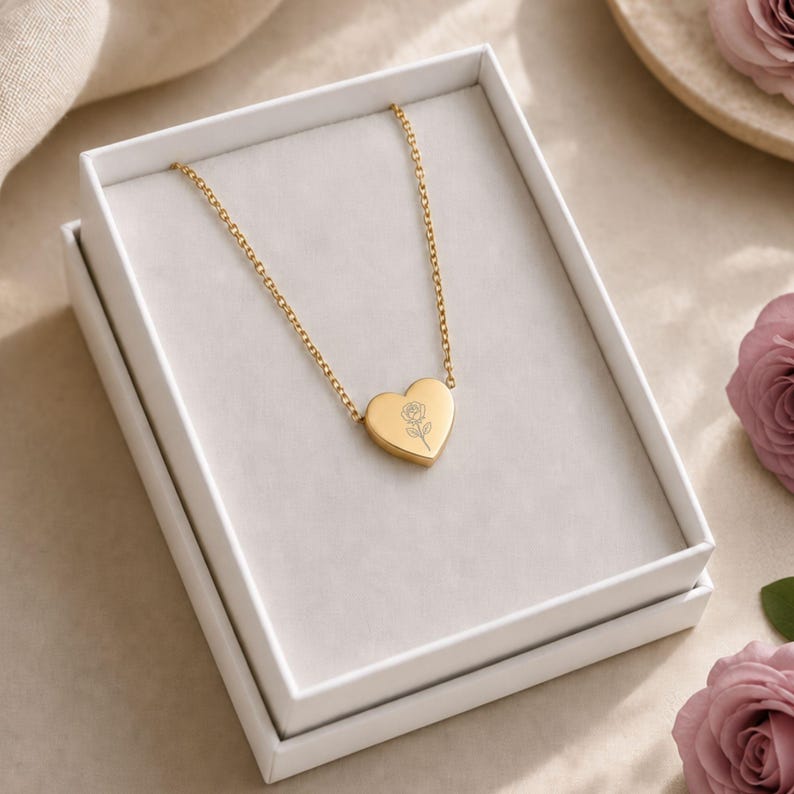 May include: A gold-toned necklace featuring a heart-shaped pendant with a delicate engraved rose design. The necklace is presented inside a white rectangular gift box, showcasing a thin chain attached to the center of the heart charm.