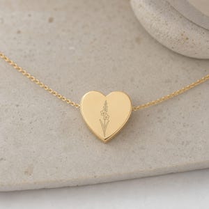 May include: A gold-toned heart-shaped pendant necklace featuring a delicate, thin chain and a central heart charm. The surface of the heart is smooth and polished, engraved with a fine-line botanical illustration of a single flower stem.