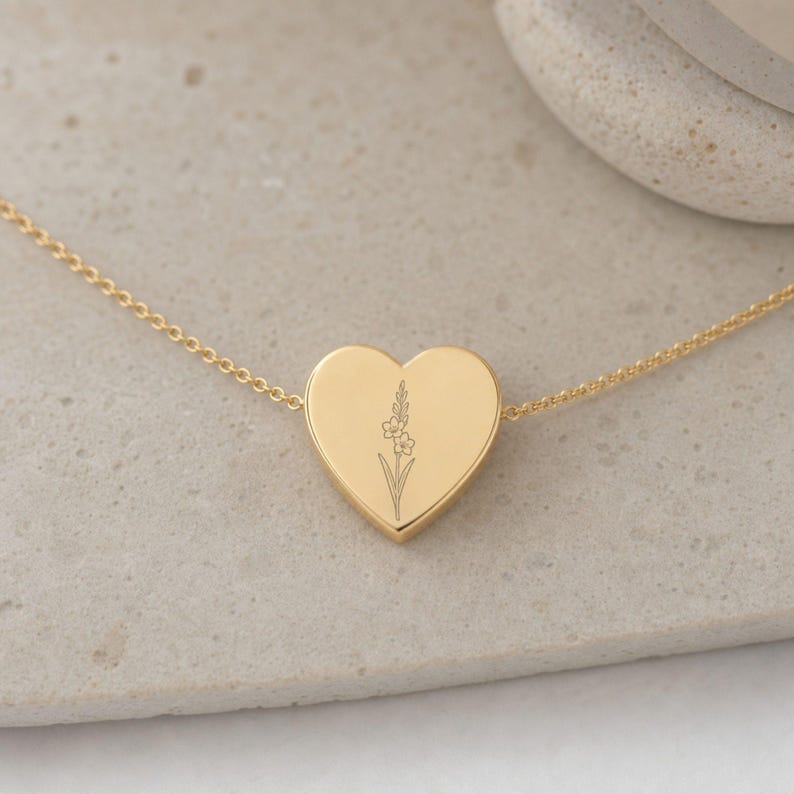 May include: A gold-toned heart-shaped pendant necklace featuring a delicate, thin chain and a central heart charm. The surface of the heart is smooth and polished, engraved with a fine-line botanical illustration of a single flower stem.