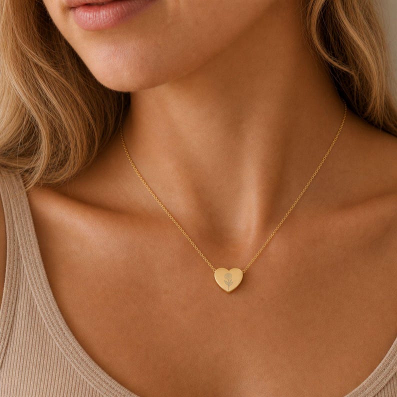 May include: A gold-toned heart-shaped pendant necklace featuring a delicate engraved flower design at the center. The pendant is suspended from a thin, matching gold-toned chain that rests against the collarbone, creating a simple and refined look.
