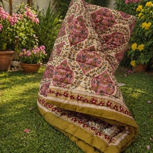 Peut inclure: Une couverture matelassée pliée avec un motif floral dans les tons de rose, de vert et d'or sur un fond crème. La couverture est posée sur de l'herbe verte, avec une bordure de motifs floraux dorés et rouges.