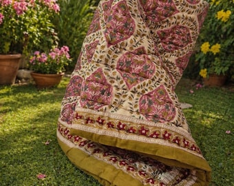 Puede incluir: Una manta acolchada y plegada con un estampado floral en tonos rosa, verde y dorado sobre un fondo crema. La manta está sobre hierba verde, con un borde de diseños florales dorados y rojos.