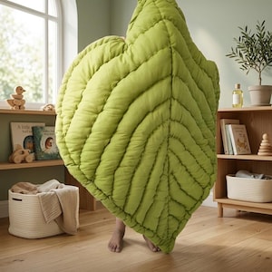 Puede incluir: Una manta grande acolchada en forma de hoja de color verde claro con costuras visibles que imitan las venas de una hoja. El texto "DESIGNED FOR NATURE'S LITTLEST ADVENTURERS" est&aacute; impreso en blanco en la parte inferior.