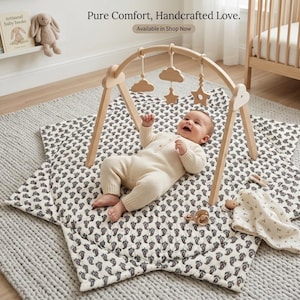 Pode incluir: Um ginásio de atividades em madeira com ornamentos suspensos de nuvens e estrelas sobre um tapete de brincar em forma de estrela com padrão paisley preto e branco. O texto Pure Comfort, Handcrafted Love. Available in Shop Now é visível.