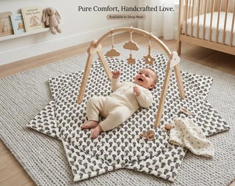 Puede incluir: Un gimnasio de actividades de madera con adornos colgantes de nubes y estrellas sobre una alfombra de juego en forma de estrella con un patrón de cachemira en blanco y negro. El texto Pure Comfort, Handcrafted Love. Available in Shop Now es visible.