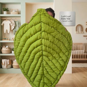Puede incluir: Una alfombra grande y acolchada para beb&eacute; con forma de hoja en un vibrante color verde lima. La alfombra presenta nervaduras cosidas detalladas que irradian desde un eje central. Un letrero en la pared dice: Lush Leaf Baby Mat - Natural Comfort.