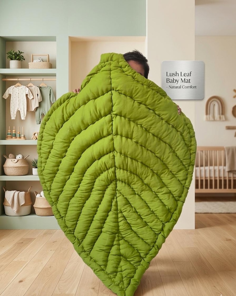 Puede incluir: Una alfombra grande y acolchada para beb&eacute; con forma de hoja en un vibrante color verde lima. La alfombra presenta nervaduras cosidas detalladas que irradian desde un eje central. Un letrero en la pared dice: Lush Leaf Baby Mat - Natural Comfort.