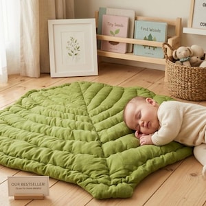 Puede incluir: Una alfombra de suelo grande con forma de hoja en color verde oliva s&oacute;lido con detalles de costuras acolchadas que imitan las venas de una hoja. La alfombra es de tela y est&aacute; colocada sobre un suelo de madera clara, con un letrero que dice OUR BESTSELLER!