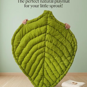 Puede incluir: Una alfombra de juego verde con forma de hoja y detalles de costuras acolchadas. El texto en la parte superior dice: ECO-FRIENDLY & PLUSH: The perfect natural playmat for your little sprout! Una pila de mantas dobladas con anillos de madera.