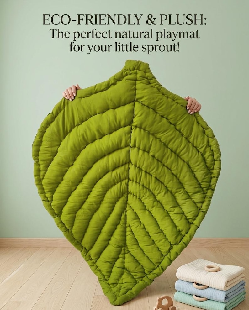Puede incluir: Una alfombra de juego verde con forma de hoja y detalles de costuras acolchadas. El texto en la parte superior dice: ECO-FRIENDLY & PLUSH: The perfect natural playmat for your little sprout! Una pila de mantas dobladas con anillos de madera.