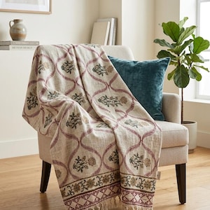May include: A cream-coloured woven throw blanket featuring a repeating lattice pattern in muted red, with small floral motifs in green and gold inside each section. The blanket has a decorative border with a geometric design and knotted fringe edges.