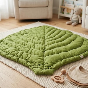 Puede incluir: Una alfombra de suelo grande con forma de hoja en un color verde vibrante. La alfombra presenta costuras acolchadas detalladas que imitan las venas de una hoja, creando una superficie texturizada y acolchada. Est&aacute; hecha de tela suave.